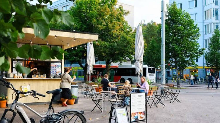 Ulkokahvila, jossa muutama asiakas istuu pöydissä puiden ympäröimällä aukiolla kaupungin keskustassa. Ulkokahvila, jossa muutama asiakas istuu pöydissä puiden ympäröimällä aukiolla kaupungin keskustassa.