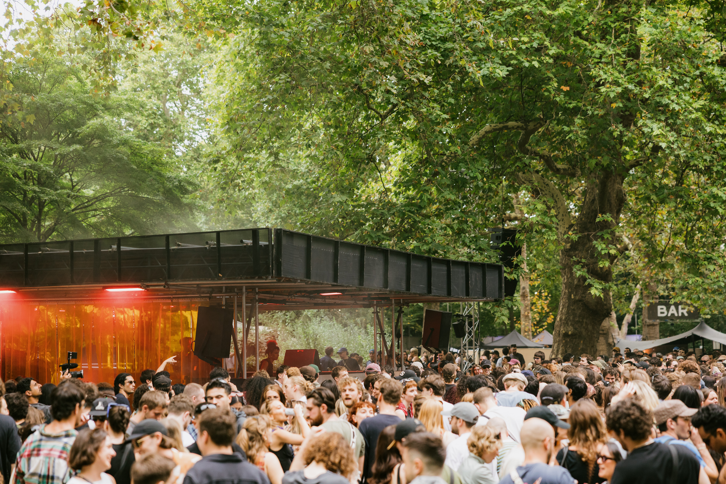 Photo of people dancing at an outdoor festival surrounded by green trees and a black stage with lights and speakers