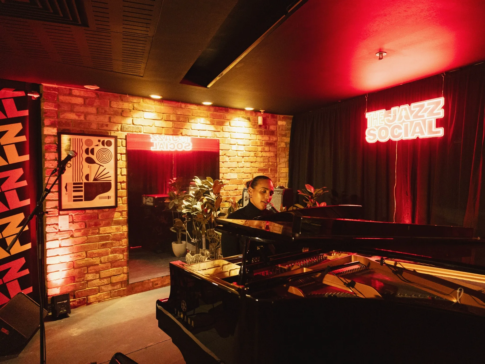 A jazz pianist playing a grand piano in front of a neon red sign saying The Jazz Social