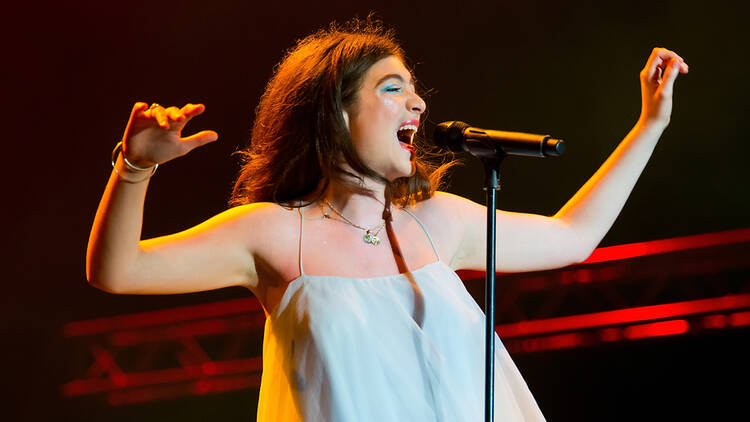 Photo of Lorde singing into a mic on stage waring a white strap dress