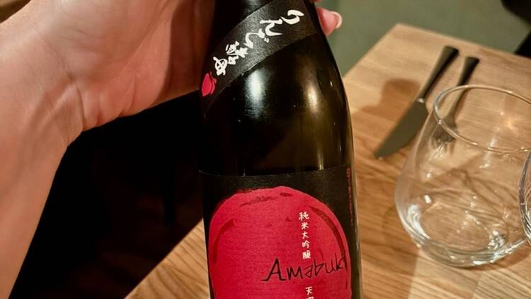 Amabuki Junmai Daiginjo -sakepullo japanilaisessa ravintolassa puupöydällä, vieressä juomalasi ja aterimet.