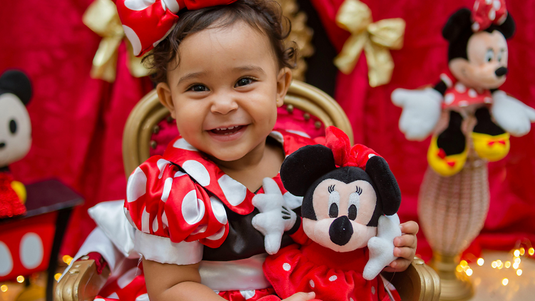 Toddler in a red and white polka dot dress holding a minnie mouse doll.