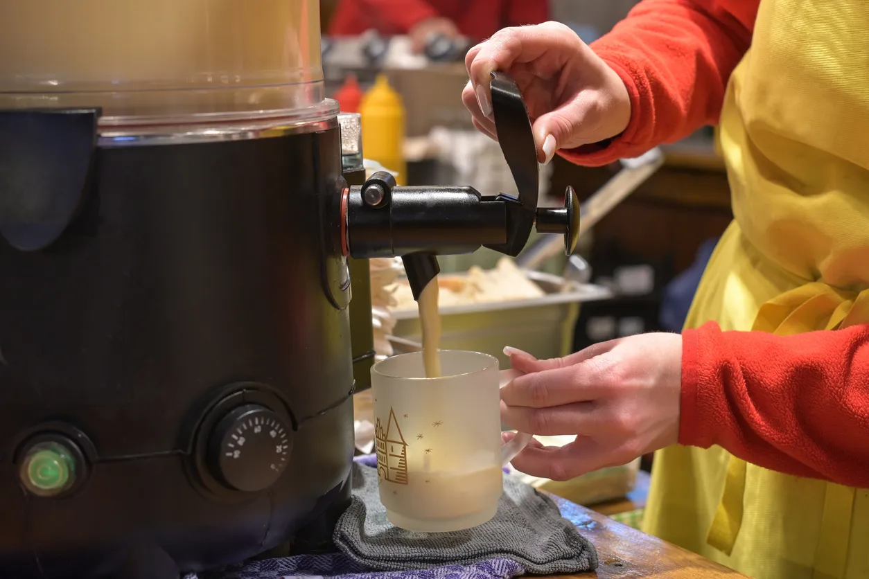 Hands of a young woman pouring warm eggnog from a cooker into a glass mug on the Christmas market, traditional hot drink with alcohol on winter holidays, selected focus