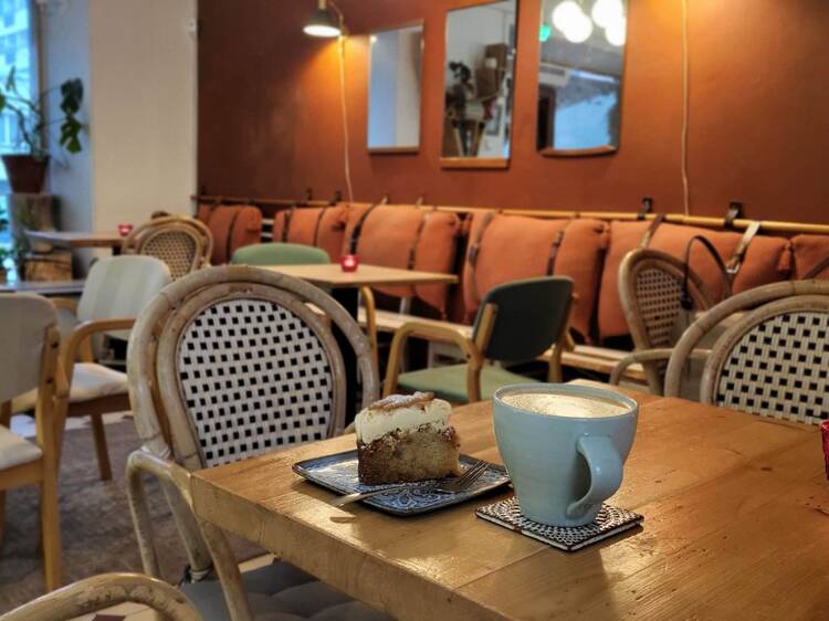 Latte ja kakkupala puupöydällä tunnelmallisessa turkulaisessa kahvilassa, jossa on boheemi sisustus ja rottinkituolit.