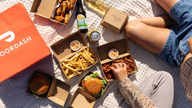 A spread of food on a picnic blanket beside a Door Dash bag 