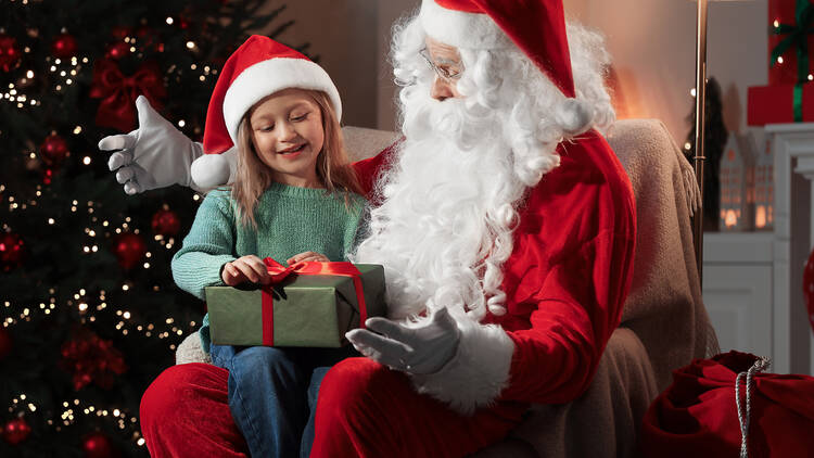 Merry Christmas. Little girl sitting on Santa's knee and holding gift at home