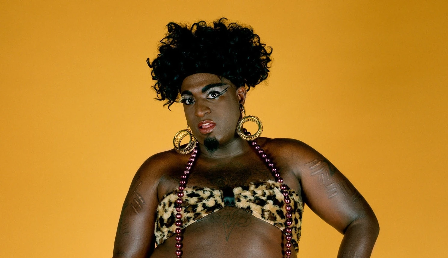 Portrait by Catherine Opie of a Black man in drag wearing a leopard print bikini in front of a yellow backdrop  