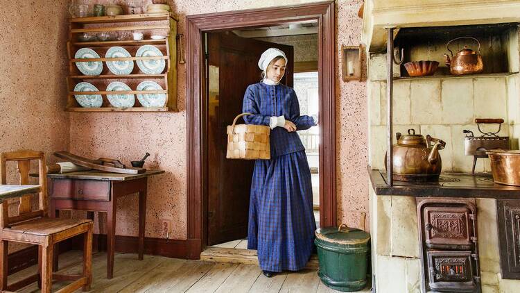 1800-luvun asuun pukeutunut nainen seisoo tuohesta tehdyn korin kanssa Ruiskumestarin talon sisällä Helsingissä