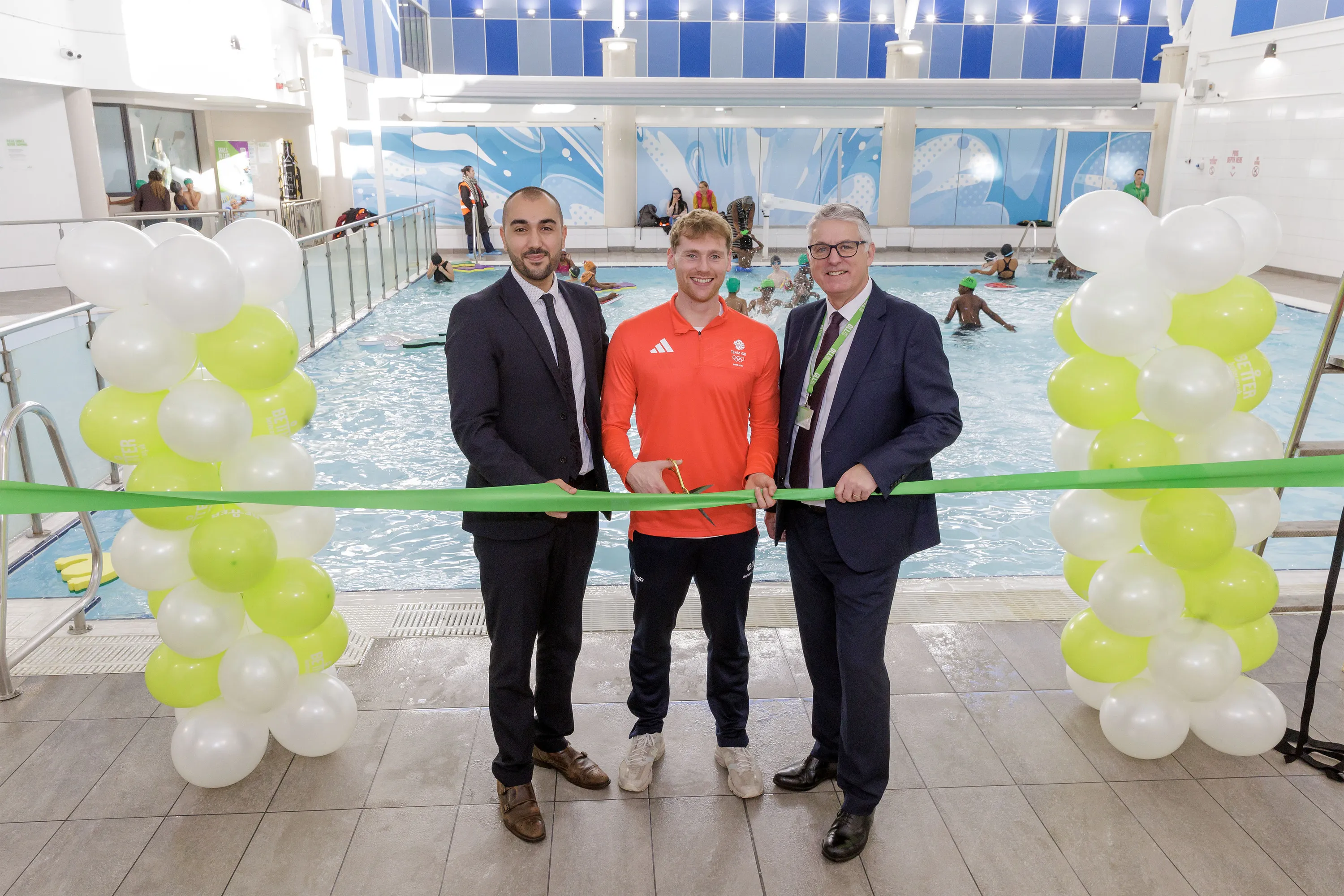 Photo of three men by a ribbon by an indoor swimming pool with baloons. One of the men is cutting the ribbon with scissors
