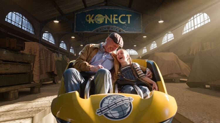 Vaari ja lapsenlapsi istuvat huvipiustolaitteen vaunun kyydissä varastohallin sisällä Vaari ja lapsenlapsi istuvat huvipiustolaitteen vaunun kyydissä varastohallin sisällä