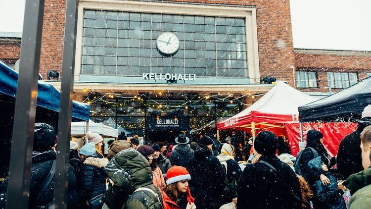 A festive crowd gathers under twinkling lights at Teurastamo’s Kellohalli entrance during a snowy Christmas market.