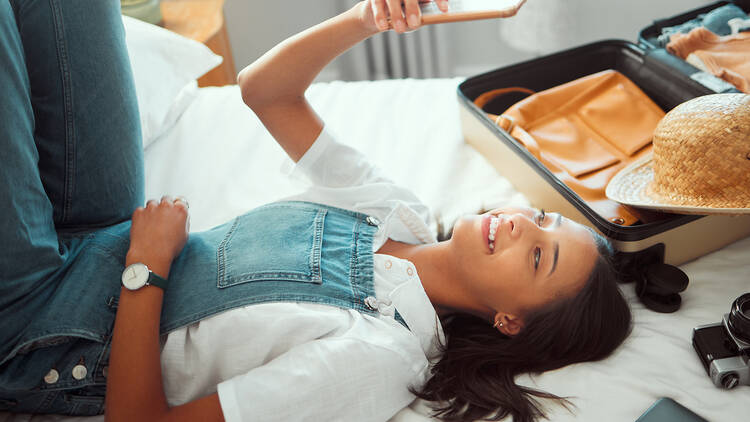 1470244255 Selfie, vacation and relax with a woman lying on a hotel bed with luggage after unpacking on vacation. Social media, phone and bedroom with a young female enjoying travel or hospitality as a tourist