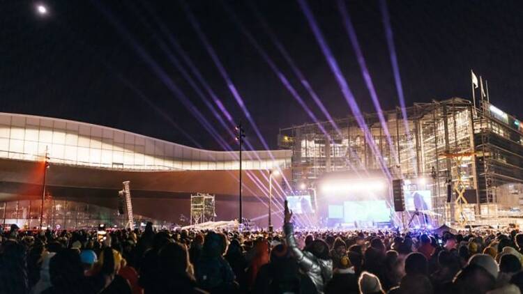 Suuri yleisö juhlii uudenvuoden konserttia Kansalaistorilla Helsingin yössä, lavalta loistaa kirkkaat valot ja taivaalle suuntautuvat värikkäät valokeilat.