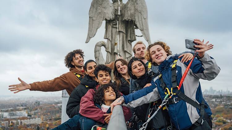A group of visitors take a selfie at the top of Summit Ally Pally, with a stone statue of an angel in the background