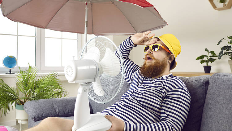 Funny sweaty chubby bearded man in swimsuit sitting at home, suffering from crazy summer heat, wiping sweat off forehead, holding electric fan, wishing for heatwave to stop and fresh breeze to blow
