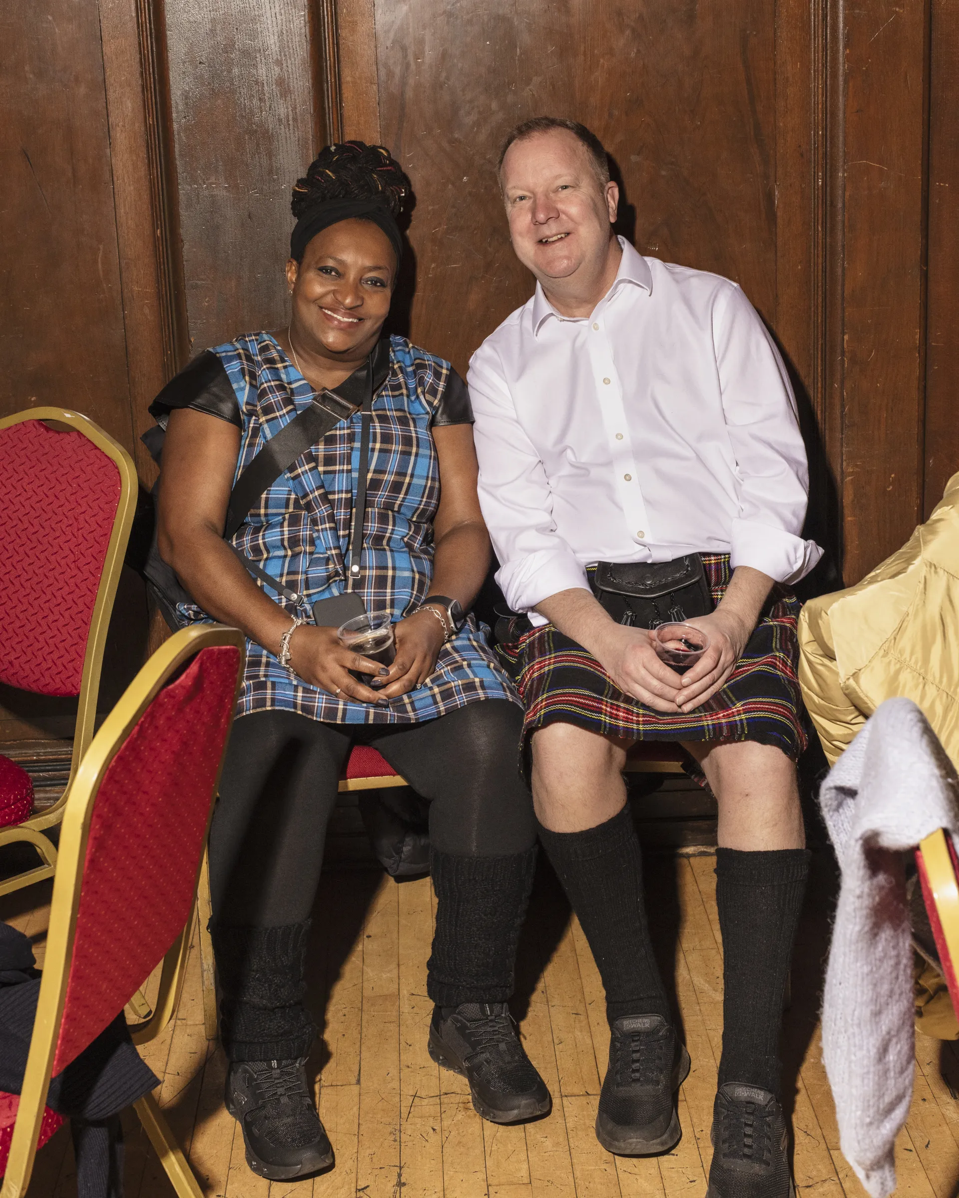 A woman and a man sat down and smiling at the camera both wearing tartan 