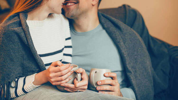 1194816164 Hands of a man and woman holding cups of hot coffee sitting on the couch at home kissing and laughing.