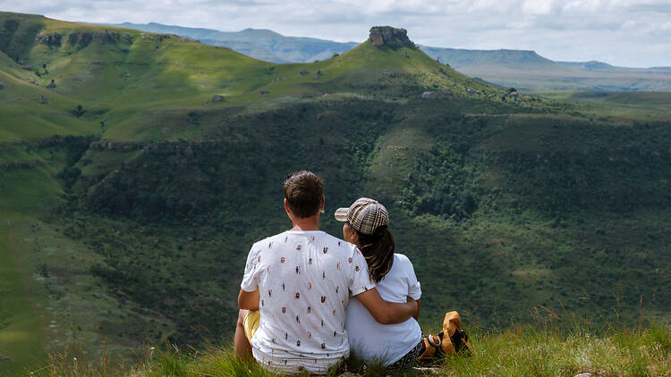 A couple relaxes in the lush greenery of Drakensberg Mountain, gazing at breathtaking landscapes. Their serene escape showcases South Africa natural beauty, perfect for travel and adventure.