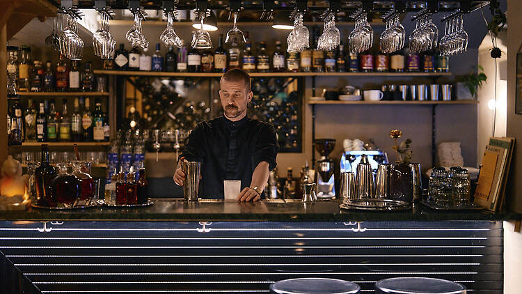 Baarimikko valmistamassa cocktailia Ravintola Kakolanruusun tunnelmallisessa baarissa, jonka hyllyillä on lukuisia pulloja ja lasipurkkeja.