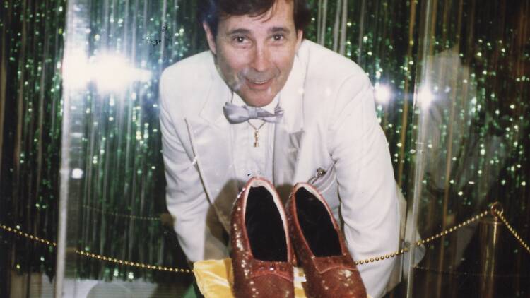 A man in a bowtie poses with the ruby slippers from The Wizard of Oz.