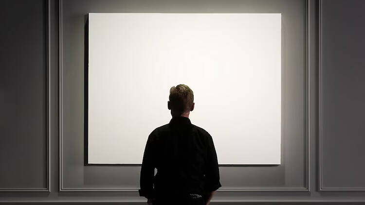 A male actor with his back to the audience, looking at an all-white picture on the wall