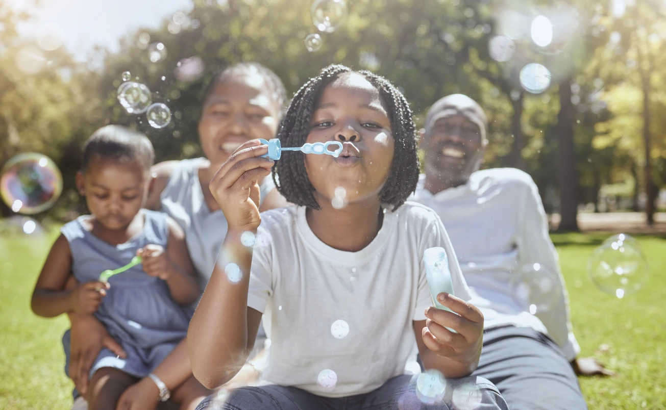 Happy, nature and black family blowing bubbles while playing, bonding and enjoying summer in the park. Happiness, father and mother with children having fun together in a green garden in South Africa