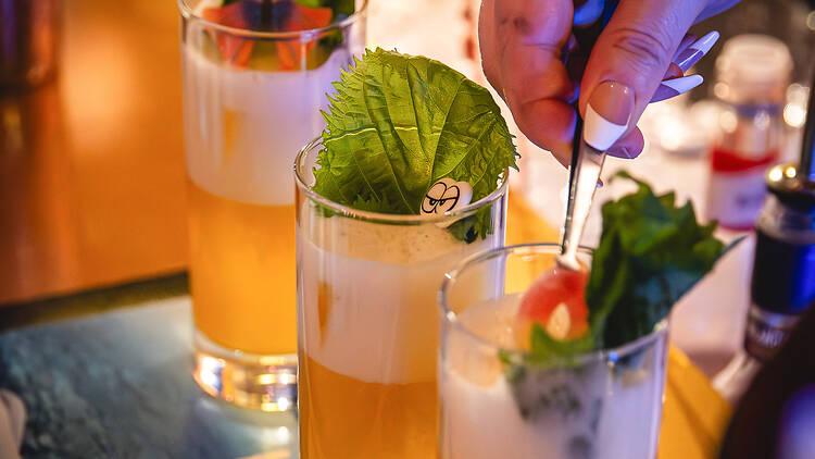 Kolme keltaista cocktailia rinnakkain ja pitkäkyntinen käsi sekoittaa yhtä niistä lusikalla