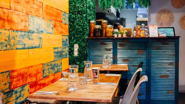 Pienikokoinen ja värikäs ravintolasali, jossa puupöydät on katettu vesikarahveilla ja ruokailuvälineillä, ja taustalla näkyy vihreää kasviseinää ja sinisävyinen tiski.