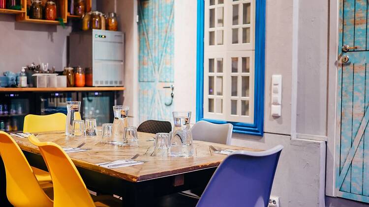 Ravintolan sisätila, jossa puupöytä on katettu vesikannuilla ja laseilla, ja ympärillä on värikkäitä tuoleja, hyllyjä sekä sinisävyisiä ovia ja yksityiskohtia.