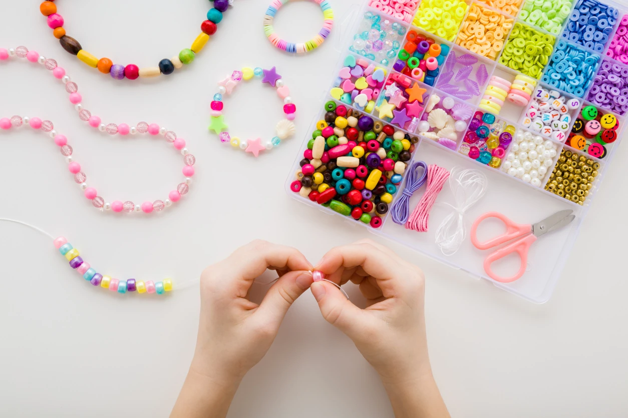 Girl hands making bracelet. Case with different colorful plastic parts on white table background. Creating beaded jewellery. Little child handcraft. Closeup. Point of view shot. Top down view.