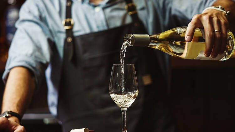 1404854443 Male sommelier pouring white wine into long-stemmed wineglasses. Waiter with bottle of alcohol beverage. Bartender at bar counter pour elite drink into long-stemmed glass
