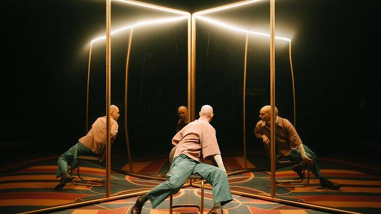 Näyttelijä istuu tuolilla peiliseinien keskellä, oma kuva ja valot heijastuvat moninkertaisina teatterilavalla.