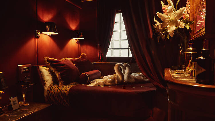 A moody, intimate bedroom with deep red walls and soft, warm lighting. A plush bed dressed in burgundy linens and textured cushions sits against the wall, with two white towel swans arranged at its centre.
