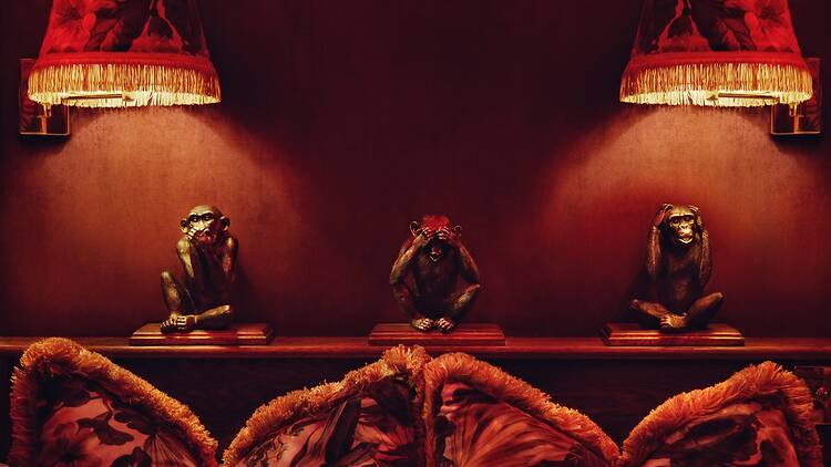 Three small monkey figurines sit beneath warm, fringed lamps on a deep red wall, evoking the “see no evil, hear no evil, speak no evil” motif above richly patterned cushions.