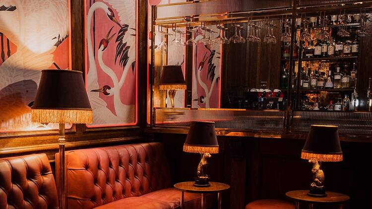 A cosy, dimly lit bar corner with tufted leather seating, fringed lamps and flamingo murals reflected in a mirrored drinks cabinet lined with glassware and bottles.
