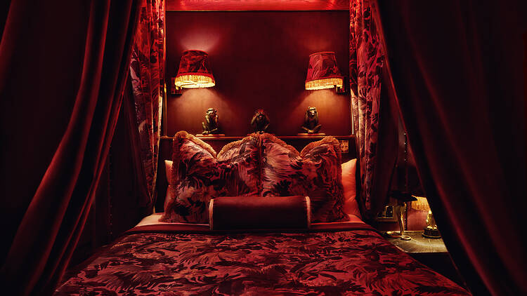 A richly decorated, red-toned bedroom with a curtained canopy bed, floral textiles, warm ambient lighting and ornamental monkey figures arranged on a shelf above plush cushions.