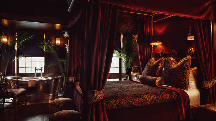 A richly decorated, red-toned bedroom with a curtained canopy bed, floral textiles, warm ambient lighting and ornamental monkey figures arranged on a shelf above plush cushions.