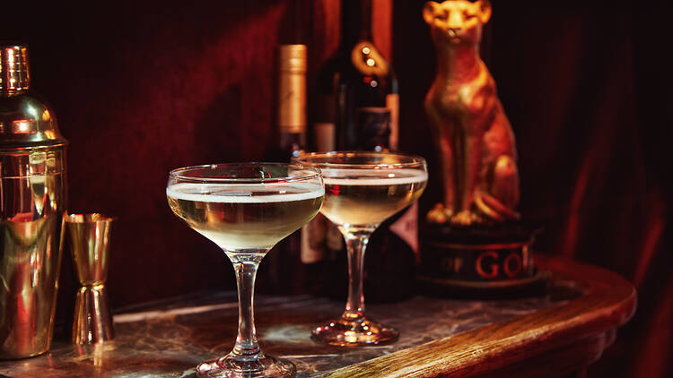 Two coupe glasses filled with a pale cocktail sit on a polished bar beside a shaker, bottles and a gold-toned leopard lamp in warm, low lighting.