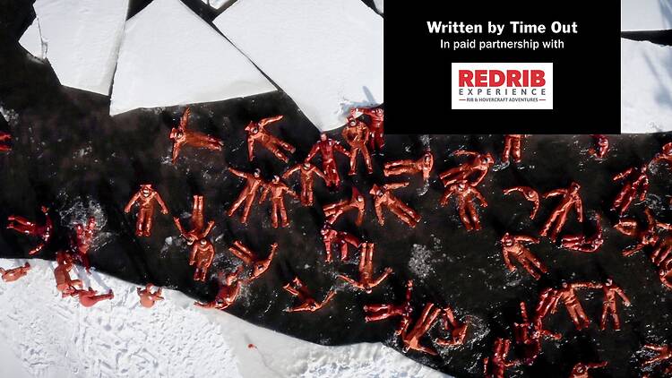 Group of people in bright orange survival suits floating in icy water during a winter ice swimming event in Helsinki archipelago.