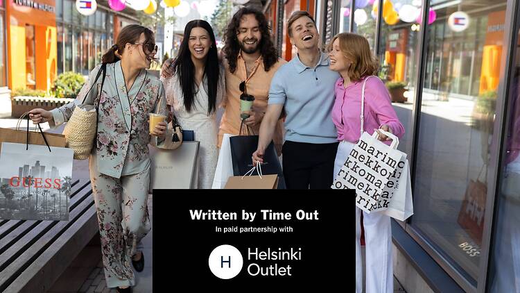 Ulkoilmakäytävällä Helsinki Outletissa kulkee joukko ihmisiä ostoskassien kanssa liikkeiden ja värikkäiden koristeiden keskellä. Ulkoilmakäytävällä Helsinki Outletissa kulkee joukko ihmisiä ostoskassien kanssa liikkeiden ja värikkäiden koristeiden keskellä.