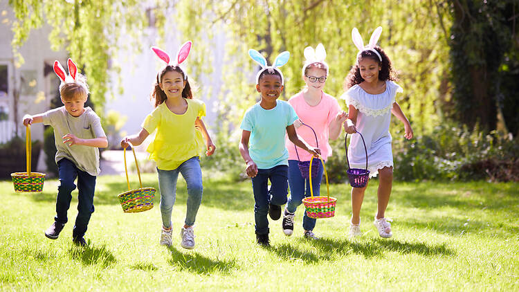 Group Of Children Wearing Bunny Ears Running To Pick Up Chocolate Egg On Easter Egg Hunt In Garden