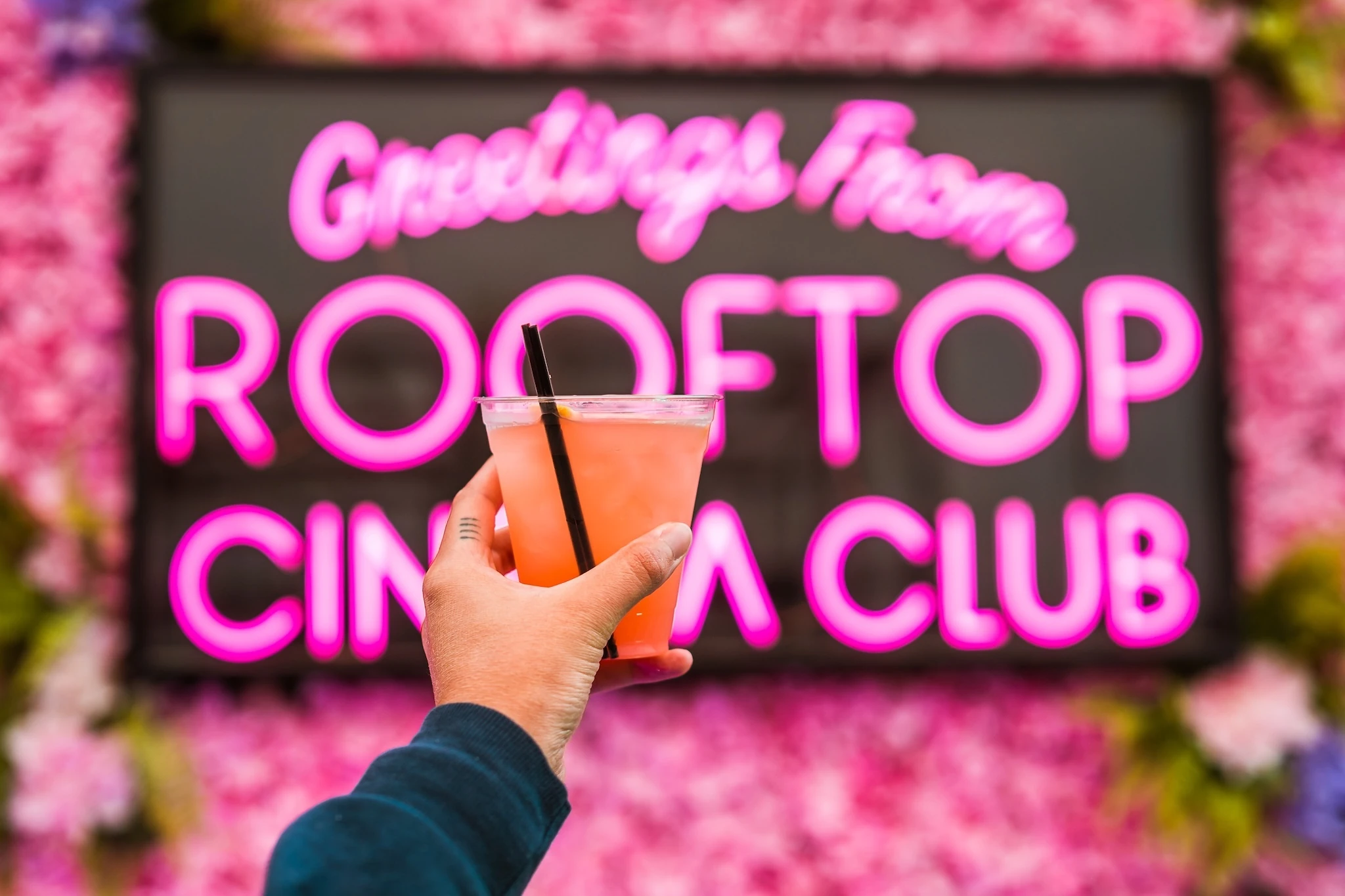 A hand raises a drink in front of a neon sign that says "Greetings from Rooftop Cinema Club"