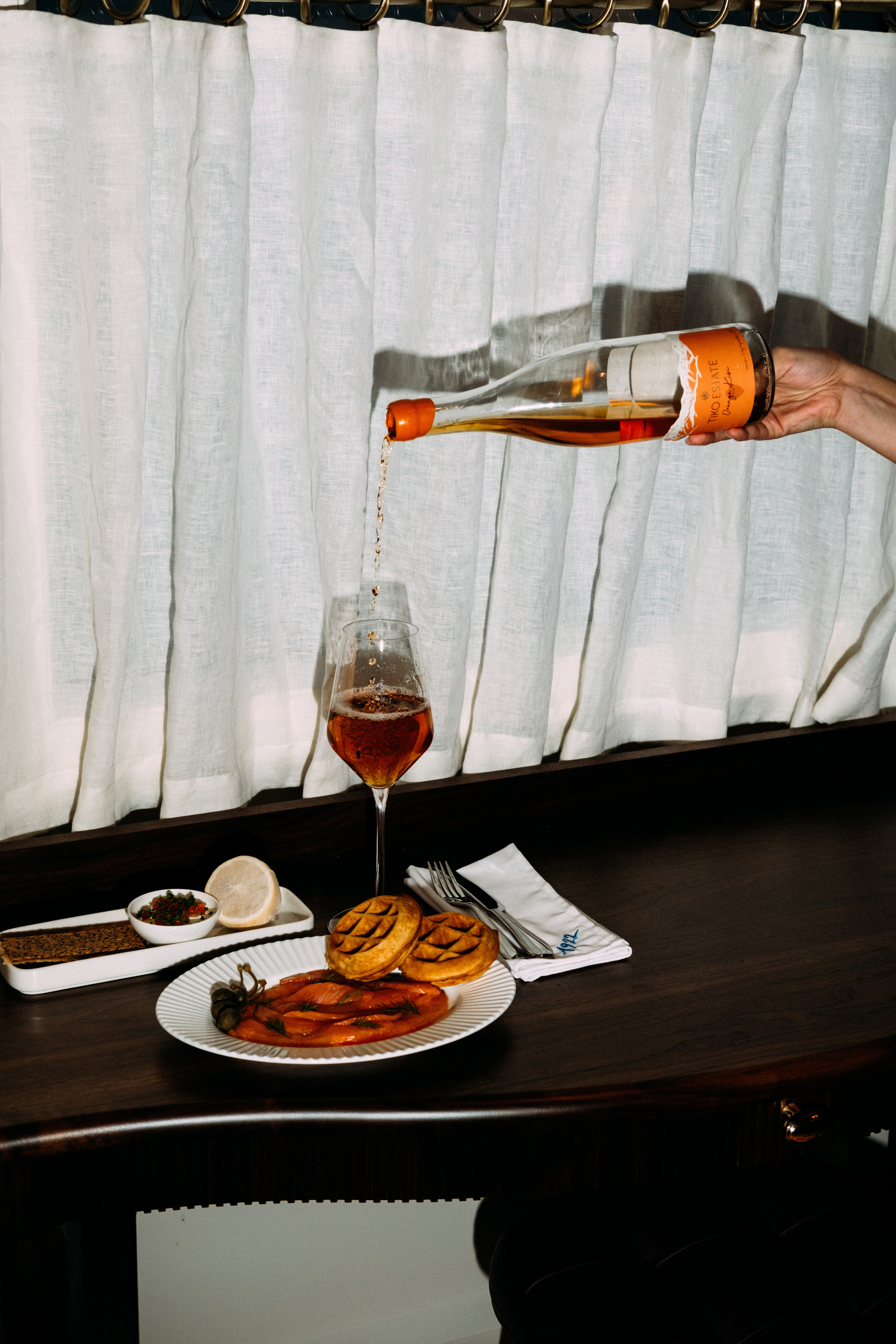 Hand pouring orange wine into glass with plate of food