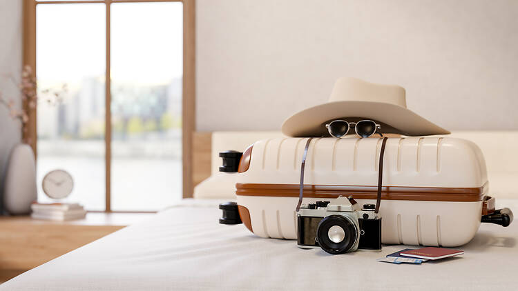 Camera and passport with sunglasses and tourist hat over beige luggage suitcase on bed. Go on a trip