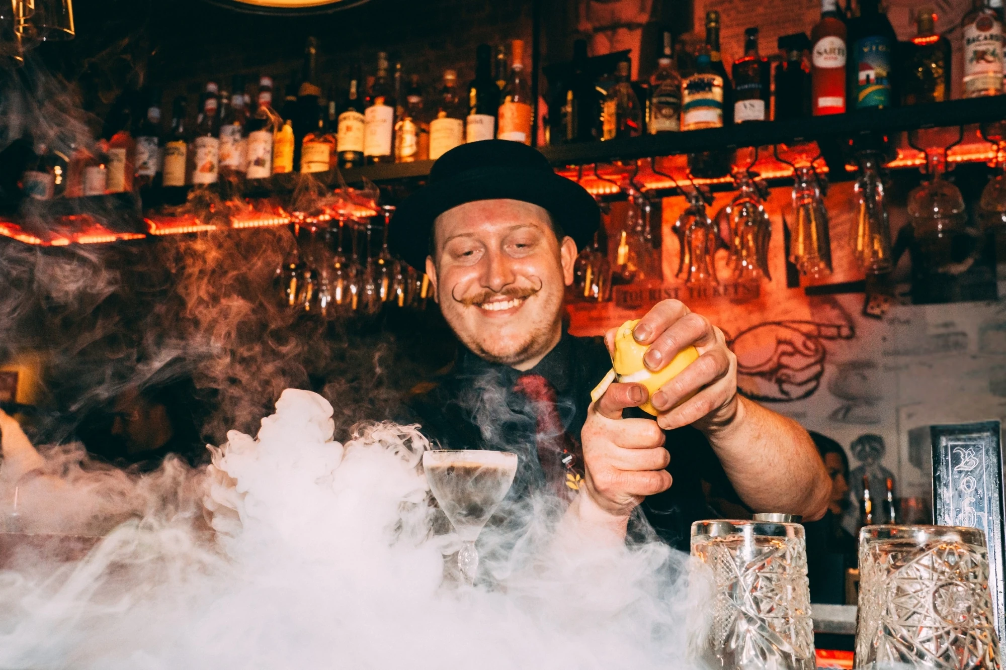 Bartender in top hat and twirly moustache creates cocktail with smoke rising from bar