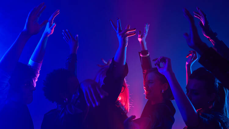1387279257 Minimal shot of diverse crowd dancing in club with hands up lit by neon lights