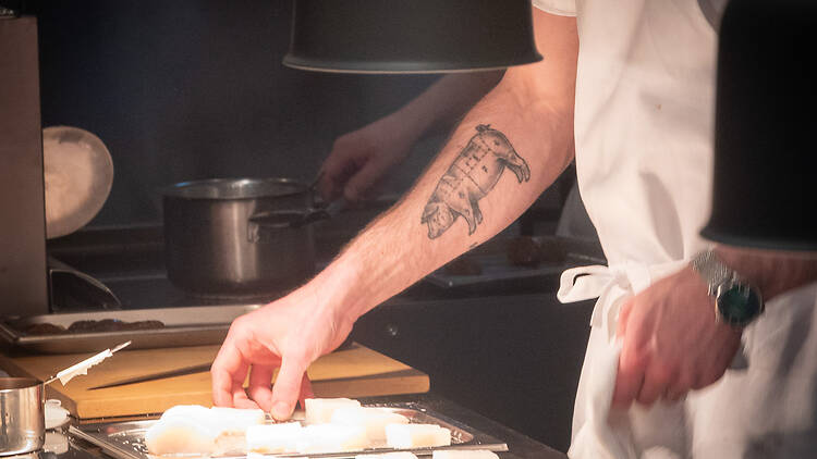 kokki asettelee kalanpalasia tarjottimelle kädessään tatuointi joka kuvaa sian eri lihanosia