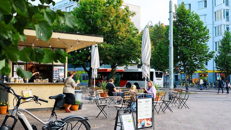 **Alt-teksti:**   Munkkiniemessä sijaitseva lippakioski, jonka edustalla on ulkopöytiä, asiakkaita istumassa, polkupyörä pysäköitynä ja taustalla kaupunkikatua busseineen ja kerrostaloineen vehreässä ympäristössä.