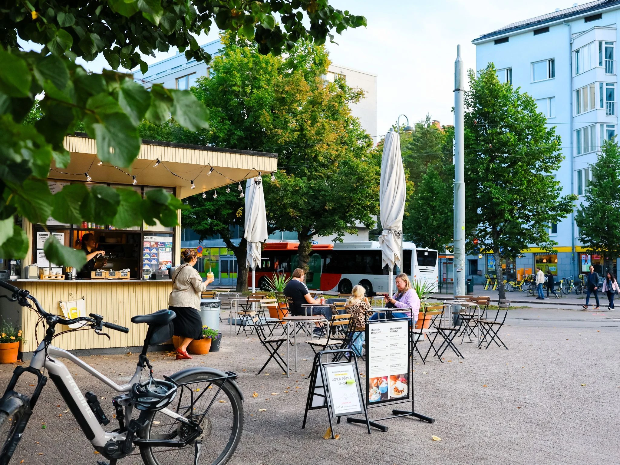 **Alt-teksti:**   Munkkiniemess&auml; sijaitseva lippakioski, jonka edustalla on ulkop&ouml;yti&auml;, asiakkaita istumassa, polkupy&ouml;r&auml; pys&auml;k&ouml;ityn&auml; ja taustalla kaupunkikatua busseineen ja kerrostaloineen vehre&auml;ss&auml; ymp&auml;rist&ouml;ss&auml;.