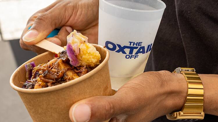 A person holding a cup of food and a glass that says "The Oxtail Off"